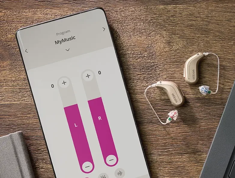 Celular com o aplicativo da Oticon aberto no controle de volume dos dois lados, ao lado de dois aparelhos auditivos pequenos