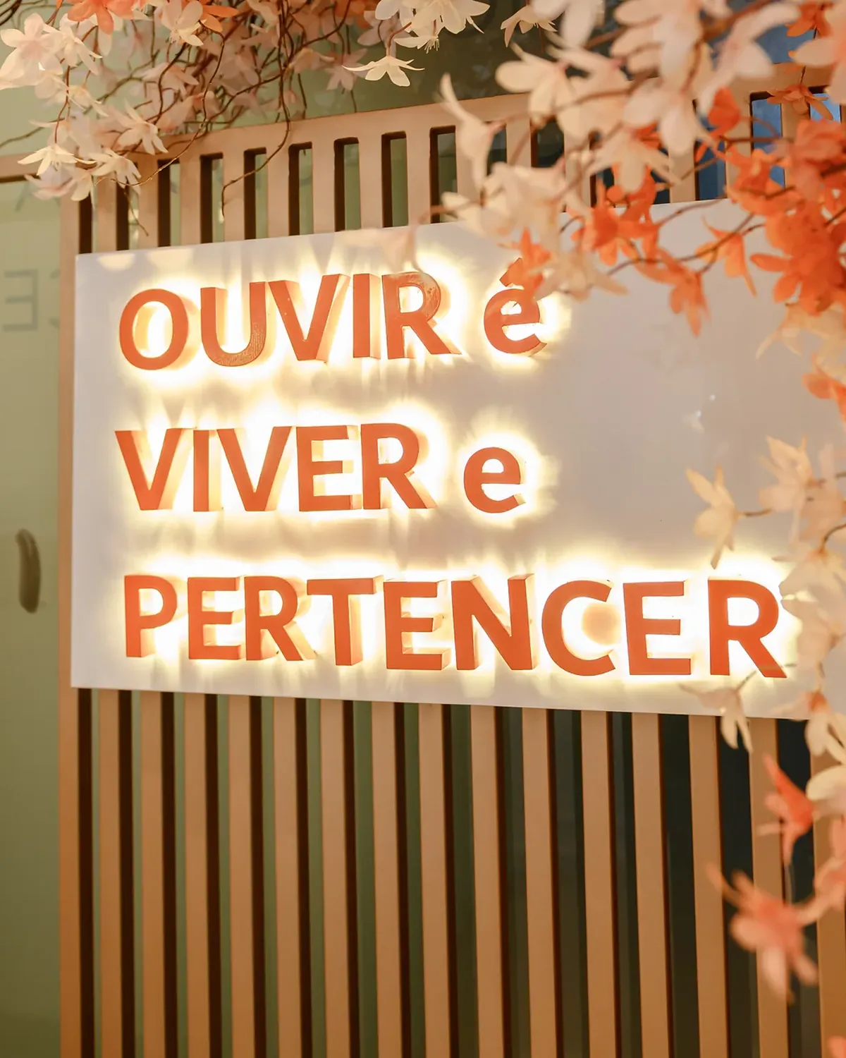 Letreiro iluminado da Hear Clean com a frase Ouvir é viver e pertencer, entre flores
