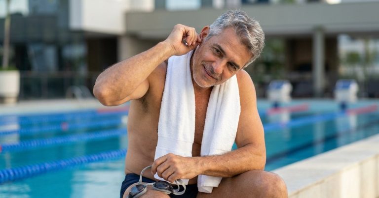 Homem de cerca de 55 anos, grisalho, sentado na borda de uma piscina com uma toalha branca no pescoço e óculos de natação na mão, inclinando a cabeça e puxando a orelha
