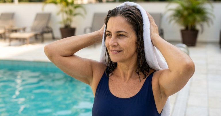 Mulher de cerca de 60 anos, de maiô azul-marinho, secando o cabelo molhado com uma toalha branca na beira da piscina de um clube