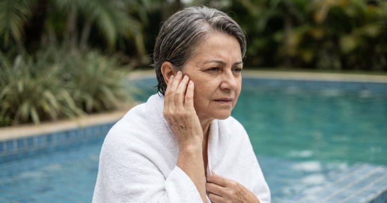 Mulher de cerca de 60 anos, de cabelo grisalho e enrolada numa toalha branca, na beira da piscina, com a mão sobre a orelha e expressão de incômodo