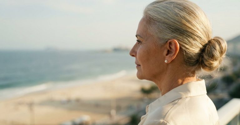 Mulher de cerca de 60 anos, de perfil, com o cabelo preso e a orelha à mostra, olhando o mar da varanda de um apartamento