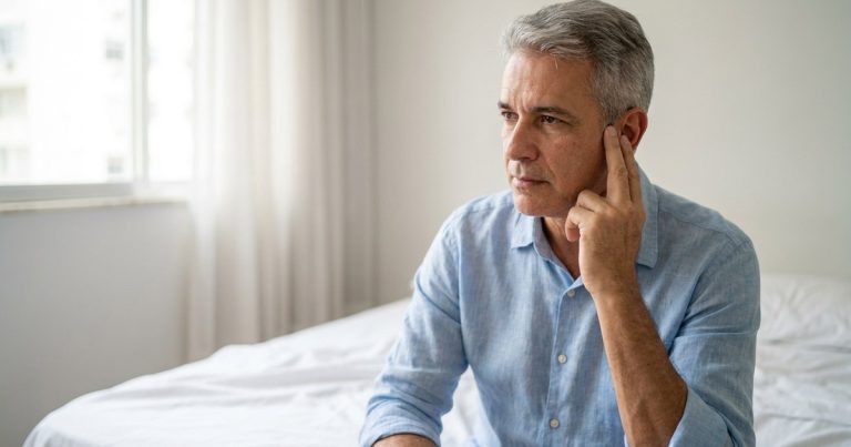 Homem grisalho de camisa azul sentado na cama pela manhã, com os dedos junto à orelha e expressão preocupada