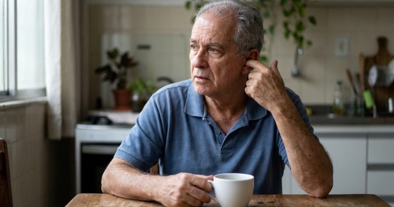 Homem de cerca de 65 anos, de cabelos grisalhos, sentado à mesa da cozinha com um dedo encostado na orelha e o olhar distante