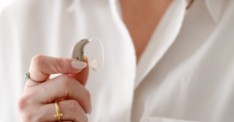 Mão da fonoaudióloga segurando um aparelho auditivo receptor no canal, com o fio transparente e a ponta de silicone na extremidade