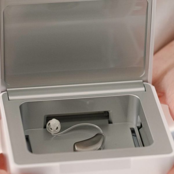Mãos da fonoaudióloga segurando um desumidificador branco aberto, com um aparelho auditivo receptor no canal guardado dentro