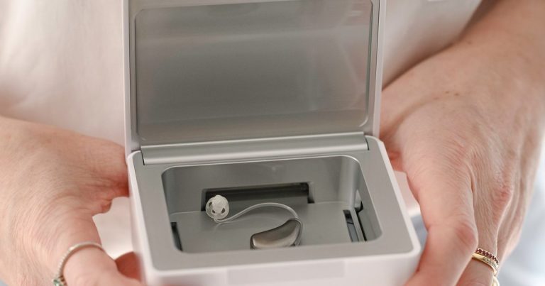 Mãos da fonoaudióloga segurando um desumidificador branco aberto, com um aparelho auditivo receptor no canal guardado dentro